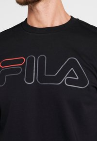 Černé bavlněné tričko s velkým obrysem loga "Fila" v šedé barvě se červeným akcentem. Design s kulatým výstřihem, krátké rukávy, hladká textura.