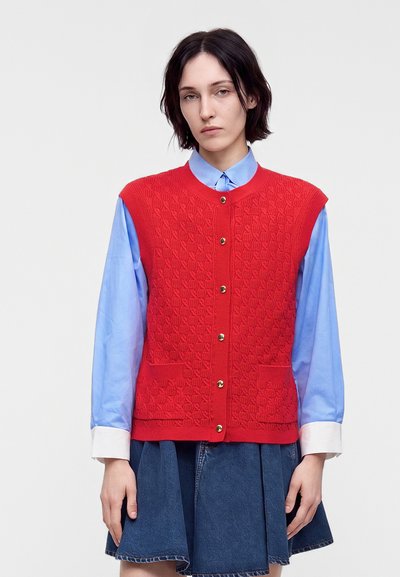 Femme portant un gilet sans manches en maille rouge boutonné sur une chemise bleu clair et une jupe plissée en denim bleu foncé, debout devant un fond uni.