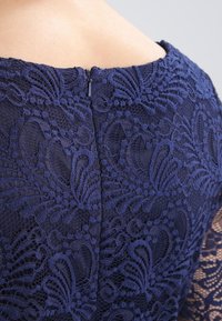 Námořnická modrá krajková šaty s detailními květinovými vzory, strukturovanou tkaninou a diskrétním zipem na zadní straně.