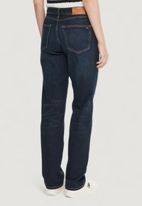 Mørkeblå denimjeans med en lige pasform, der har en brun læderlogo-patch bagpå og to baglommer. Hvide sneakers bæres.