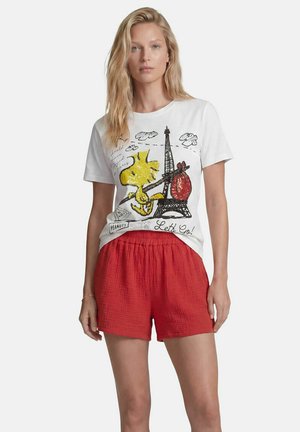 Frau trägt weißes T-Shirt mit Woodstock- und Eiffelturm-Aufdruck, kombiniert mit lockeren roten Shorts, steht vor einfachem hellem Hintergrund.
