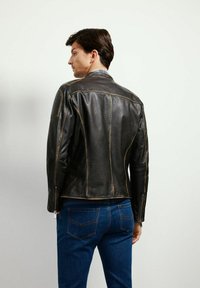 Czarna skórzana kurtka motocyklowa z ukośnymi szwami, zamkami na mankietach i kontrastowymi szwami; noszona z niebieskimi dżinsami.