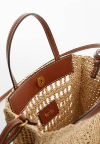 Sac en paille tressée avec des accents en cuir marron, présentant un dessus ouvert, une fermeture à bouton et une étiquette rectangulaire avec le texte en relief "MNG."
