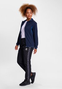 Marineblauwe hoodie met blauwe accenten, wit T-shirt, zwarte trainingsbroek met een chevronpatroon en zwarte sportschoenen.