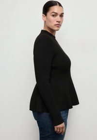 Top lungo a maniche nere a coste con colletto alto, corpo aderente e orlo svasato; abbinato a jeans in denim blu.