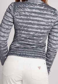 Femme portant une veste grise matelassée ajustée et un jean blanc taille haute avec un petit logo triangulaire sur la poche arrière.