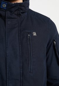 Marinblå jacka med hög krage, fickor med dragkedja och en metalllogotyp som accent. Tillverkad av ett strukturerat tyg med en slitstark finish.