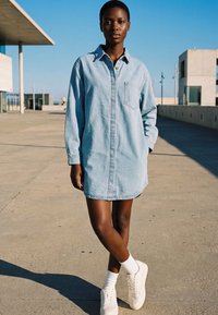 Young woman standing outdoors on concrete surface wearing light blue denim shirt dress, white socks, and white sneakers in daylight.