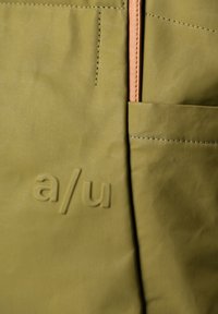 Olivegrüne Tasche aus strukturiertem Material, mit einem hellbraunen Reißverschluss und einer Vordertasche mit geprägtem "a/u"-Logo.