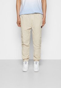 Pantalones cargo beige con cintura elástica, puños elásticos, bolsillos laterales y un pequeño parche de logo; combinados con zapatillas blancas.