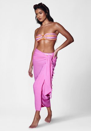 Vrouw draagt een kleurrijke bandeau bikinibovenstuk met een oranje ring en een gebonden roze sarongrok, staat op blote voeten met een zonnebril op haar hoofd.