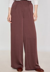 Pantaloni a gamba larga in un ricco tessuto bordeaux, caratterizzati da una texture liscia, pieghe frontali e tasche laterali. Dettagliati con una vita alta.