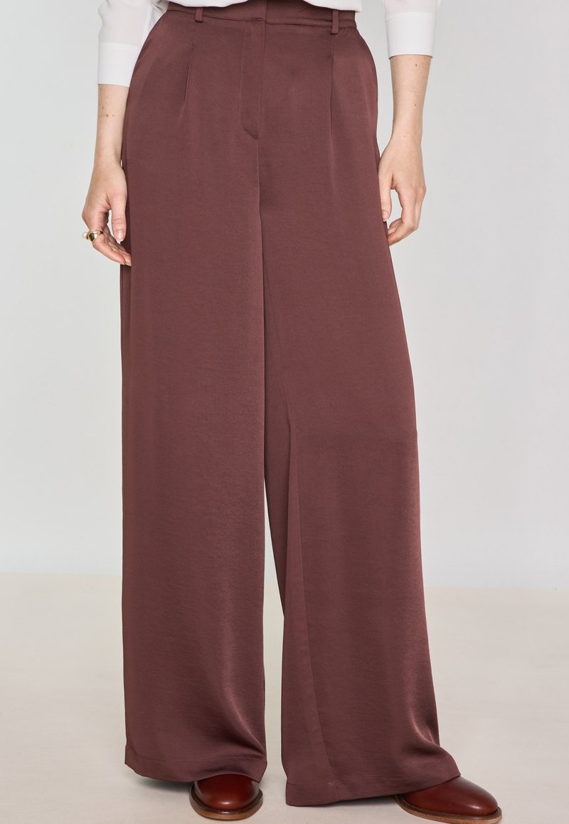 Pantaloni a gamba larga in un ricco tessuto bordeaux, caratterizzati da una texture liscia, pieghe frontali e tasche laterali. Dettagliati con una vita alta.