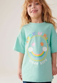 Turkis kortærmet t-shirt med et grafisk motiv af et smilende ansigt, stjerner og en regnbue, og teksten "HAPPY & KIND siden 1974".