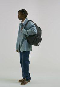 Batoh s matným černým povrchem, nošený na pravém rameni. Model v světle modré džínové bundě, tmavě modrých kalhotách a hnědých botách.