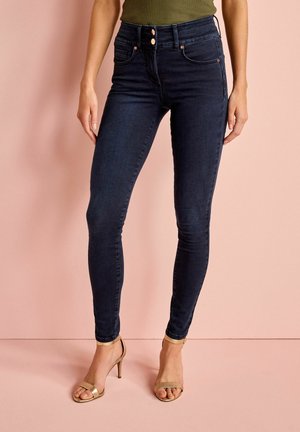 Tmavě modré, vysoké skinny džíny s měděnými knoflíky a stehy. V kombinaci s sandály na vysokém podpatku, otevřenými na prstech, v metalické zlaté barvě. Hladká denimová textura.