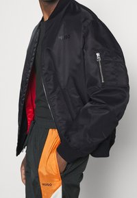 Giubbotto bomber nero con una texture liscia e lucida, polsini e colletto a costine; presenta foderatura rossa, tasca con zip sulla manica e dettaglio del logo.