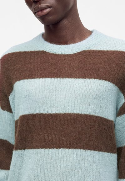 Personne portant un pull en maille douce avec de larges rayures horizontales bleu clair et marron, montré du cou au milieu du torse.