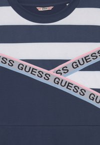 Marinblå och vit randig t-shirt med rund halsringning, med två diagonala remmar som har texten "GUESS" i svart på rosa och blå bakgrund.