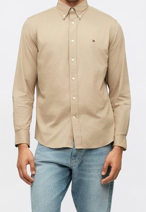 Mann trägt ein beiges Hemd mit Knöpfen und kleinem Logo auf der Brust sowie hellblaue Jeans und steht mit entspannten Händen.