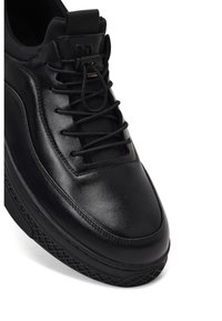 Zapatilla de cuero negro con punta redondeada, superficie texturizada y sistema de cordones. Presenta una suela de goma con patrón y un talón reforzado.