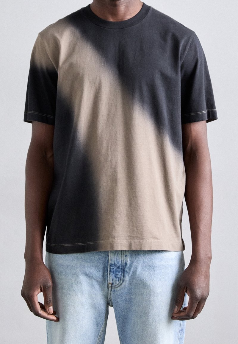 Homme portant un t-shirt à manches courtes col rond avec un motif dégradé noir et beige, associé à un jean en denim bleu clair.