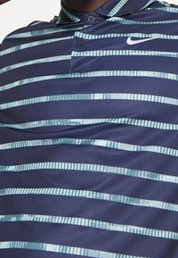 Polo shirt marine avec des rayures horizontales bleu clair, comportant un col à deux boutons et un petit logo Nike sur la poitrine. Texture de tissu douce.