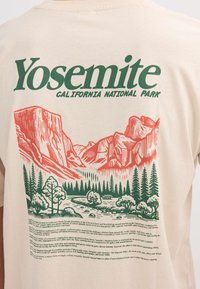 Camiseta del Parque Nacional Yosemite en California con ilustración de montaña roja y bosque verde y texto descriptivo sobre el parque en tela color crema.