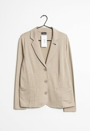 Blazer beige de tres botones con solapas con muesca y bolsillos delanteros colgado en una percha negra sobre un fondo blanco.