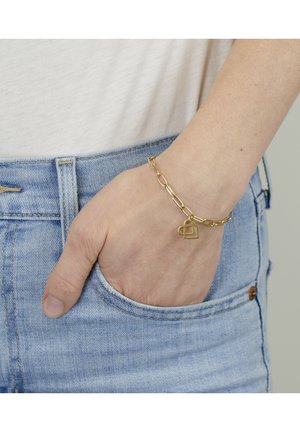 Gold chain bracelet featuring elongated links and a heart-shaped charm, worn on a hand with fingers visible in a denim pocket.