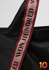Black fabric tote bag featuring a wide maroon strap with “WON HUNDRED” text. Smooth texture, square top, and visible stitching along edges.