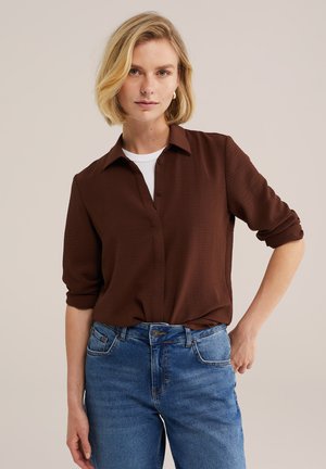 Femme blonde aux cheveux courts portant une chemise marron boutonnée sur un haut blanc et un jean bleu, debout avec une main sur la hanche.
