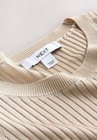 Encolure de pull en maille côtelée beige avec une étiquette blanche indiquant la marque "NEXT" et le texte "Made in China".