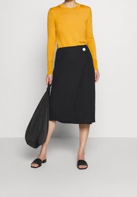 Maglione giallo a maniche lunghe abbinato a una gonna avvolgente nera con un bottone bianco, sandali piatti neri e una borsa nera.