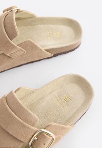 Sandales en daim beige avec une large bride ornée d'une boucle dorée, semelle intérieure rembourrée et cambrure texturée pour le confort. Semelle extérieure marron profilée basse.