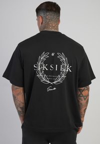 Vīrietis ar kakla un roku tetovējumiem valkā melnu pārāk lielu T-kreklu ar baltu lauvas vainagu un "SikSilk" logo uz muguras.