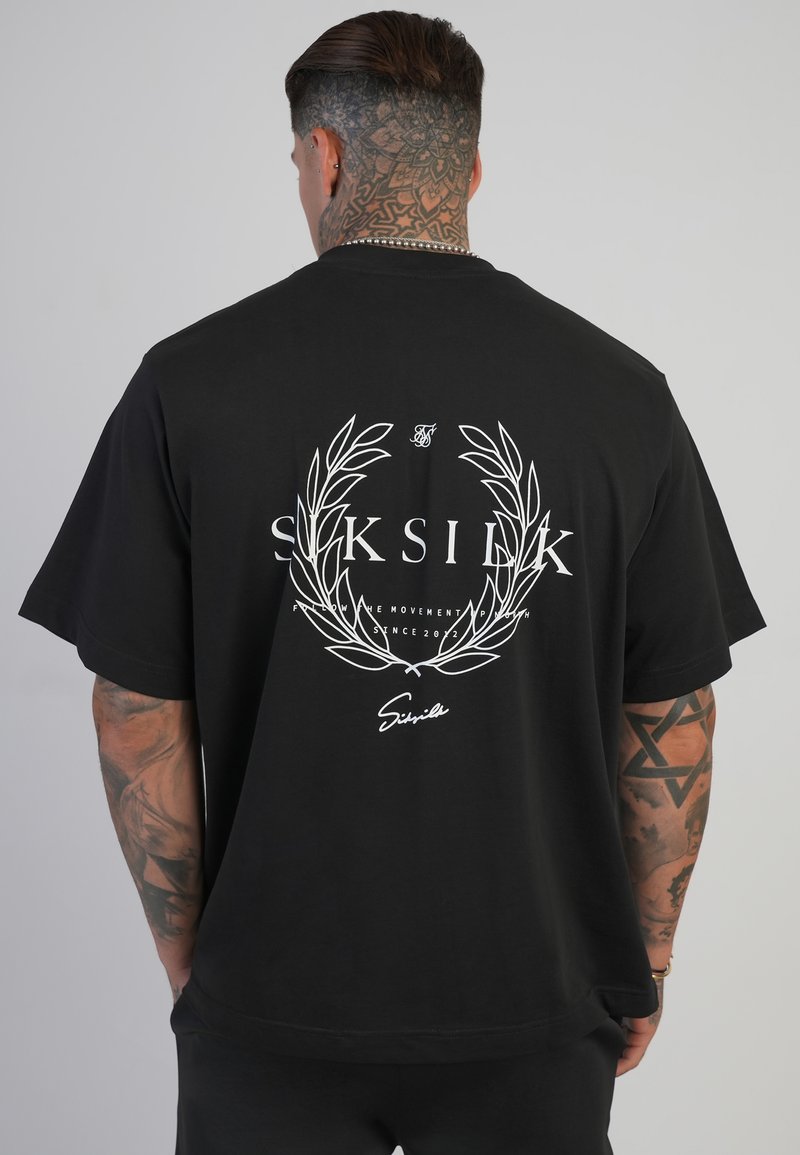 Vīrietis ar kakla un roku tetovējumiem valkā melnu pārāk lielu T-kreklu ar baltu lauvas vainagu un "SikSilk" logo uz muguras.