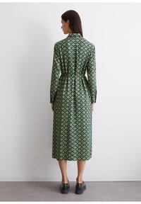 Abito lungo verde con un motivo geometrico in giallo e crema. Il design presenta un colletto, maniche lunghe e una vita cinchata.