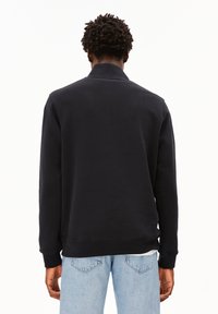 Schwarzer Pullover mit hohem Kragen, langen Ärmeln und gerippten Bündchen. Kombiniert mit hellblauen Jeans, die einen lässigen Schnitt zeigen.