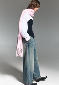Breda denimjeans, marinblå kordtopp och en rosa stickad halsduk. Halsduken har en fransad kant. Modellen står framför en neutral bakgrund.
