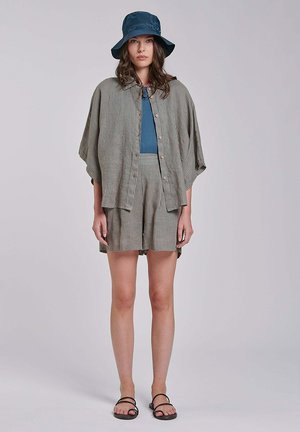 Donna con cappello a secchiello blu, top blu, camicia larga a righe con bottoni, pantaloncini abbinati e sandali neri, in piedi su sfondo grigio.