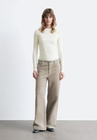 Langärmliges, hellgelbes Oberteil mit figurbetontem Design, kombiniert mit weit geschnittenen beigefarbenen Cordhosen mit feinen vertikalen Rippungen und einem Gürtel.