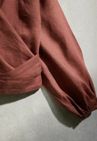 Rostfarbener langärmliger Pullover mit gesammeltem elastischem Bündchen und gefaltetem Detail nahe der Taille, präsentiert auf einer hellgrauen Oberfläche.