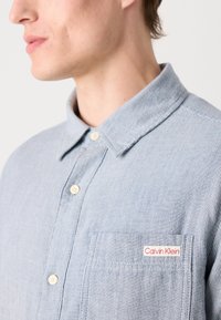 Világoskék csíkos ing, gombos gallérral, elülső zsebbel és egy kis piros "Calvin Klein" logóval a bal oldalon.