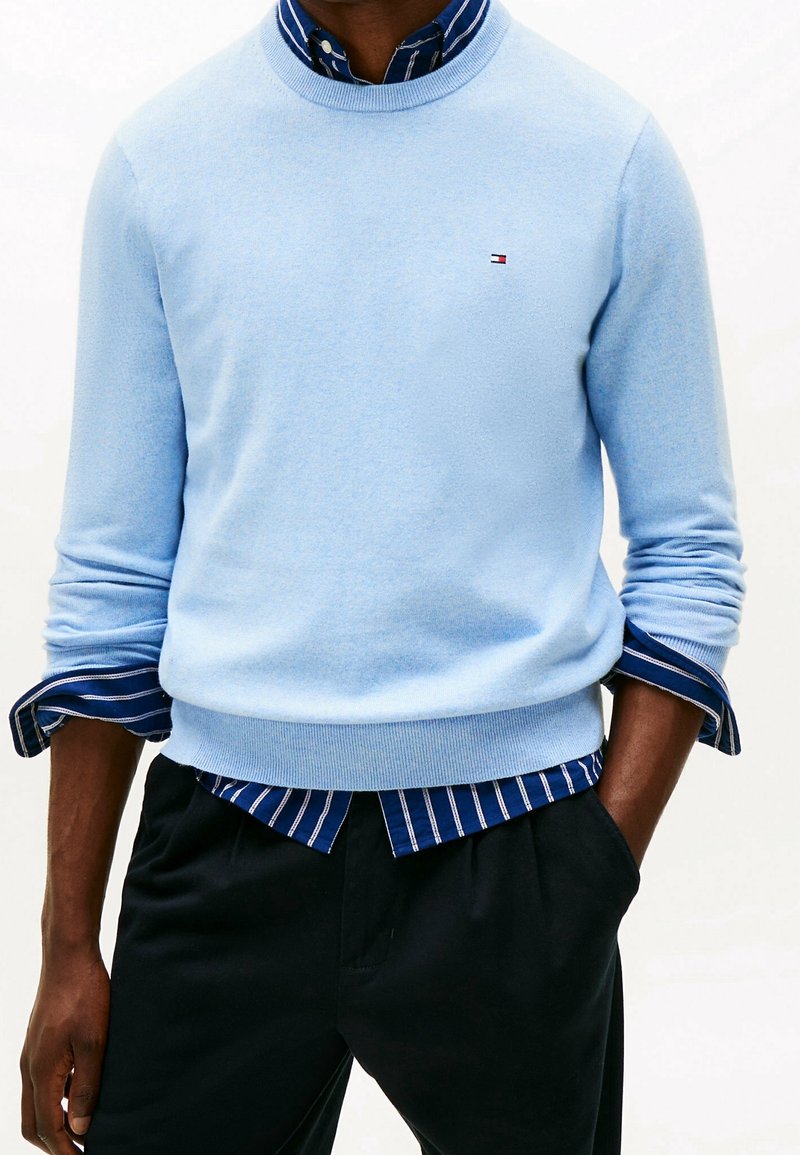 Homme portant un pull col rond bleu clair sur une chemise rayée bleue boutonnée, pantalon noir, mains dans les poches, sur fond blanc.