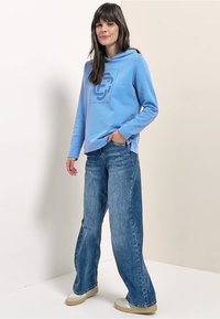 Lichtblauwe hoodie met een grafische print, gecombineerd met wijde blauwe jeans en beige schoenen. De stof oogt zacht en casual.