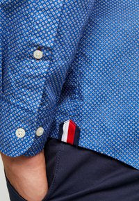 Blauw gestippeld shirt met kleine geometrische ontwerpen. Voorzien van witte knopen en een vlagdetail in rood, wit en blauw aan de zoom.