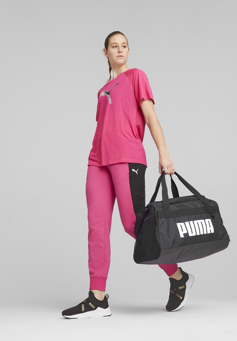 Pink short-sleeve shirt with a logo, matching pink joggers with black side panels, black sneakers with gold accents, and a black duffel bag.