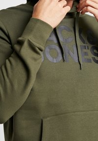 Olivgrön hoodie i mjuk bomull med framficka och dragsko på huvan, med stor svart text "BACK & JONES" tydligt över bröstet.