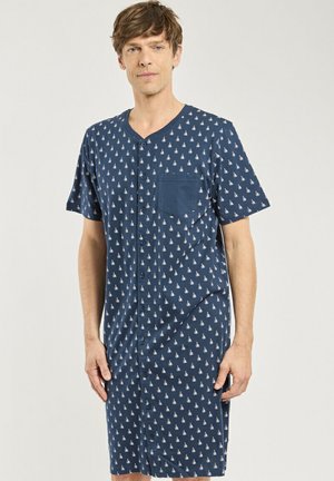 Homme portant une chemise de nuit bleue à boutons avec de petits motifs de voiliers blancs et une poche poitrine, debout devant un fond uni.